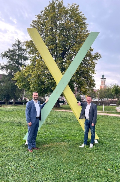 Bild: Das Bild zeigt eine Wiese mit Bäumen und einem Gebäude rechts in der Ecke. In der mitte des Bildes steht ein großes X aus Holzbalken in den Farben Gelb und Mintgrün. Dieses X stellt ein Symbol für das Tal X dar. Links von dem X ist der Oberbürgermeister von Freudenstadt Adrian Sonder abgebildet und rechts von dem X steht der Bürgermeister von Baiersbronn, Michael Ruf.